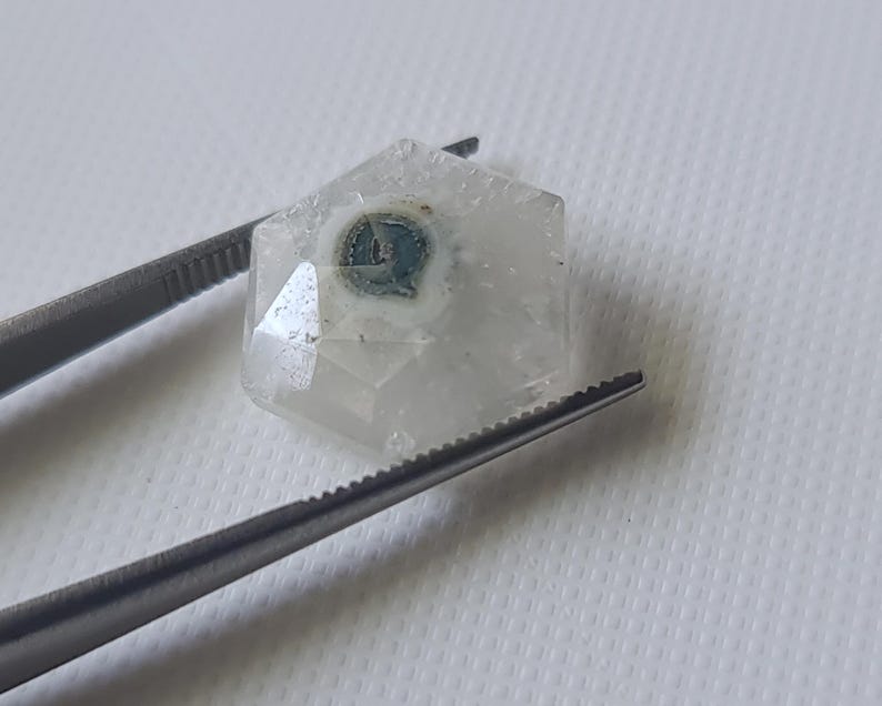 May include: A clear, faceted, hexagonal crystal with a blue and white inclusion in the center. The crystal is held by tweezers.