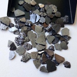 Peut inclure: Une collection de petites pierres noires et argentées de forme irrégulière. Les pierres sont éparpillées sur une surface blanche.
