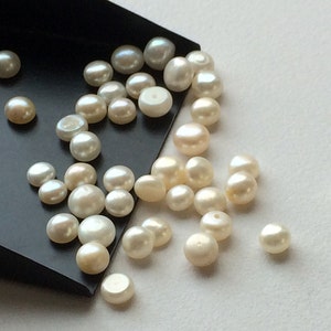May include: A collection of white round pearls of varying sizes, scattered on a white surface and a black tray.