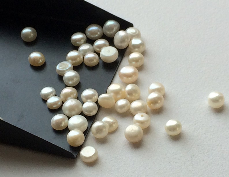 May include: A collection of white round pearls of varying sizes, scattered on a white surface and a black tray.