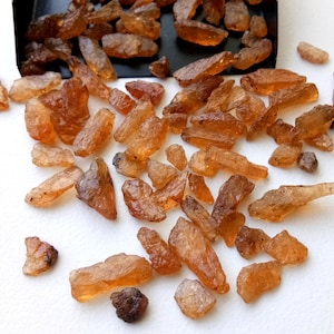 May include: A collection of small, rough, orange-brown gemstones. The gemstones are scattered on a white surface.