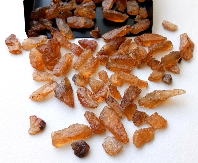 May include: A collection of small, rough, orange-brown gemstones. The gemstones are scattered on a white surface.