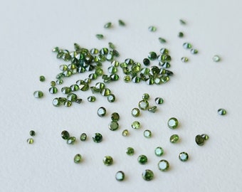 May include: A scattering of small, round, green gemstones on a white surface. The gemstones are faceted and have a shiny, reflective surface.