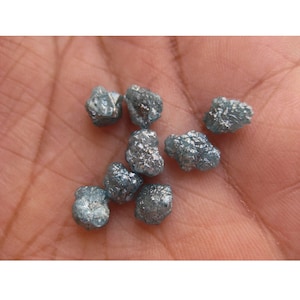 May include: A collection of small, rough, gray-blue diamonds. The diamonds are scattered on a hand.