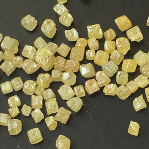 May include: A collection of small, yellow, faceted, cubic crystals. The crystals are scattered on a dark surface.
