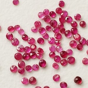 Könnte beinhalten: Eine Ansammlung kleiner, facettierter, runder Edelsteine in verschiedenen Rot- und Rosatönen. Die Edelsteine sind auf einer weißen Oberfläche verstreut, wobei sich das Licht auf ihren geschliffenen Oberflächen spiegelt. Es handelt sich um lose Rubine.