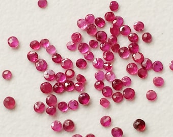 May include: A collection of small, faceted, round gemstones in varying shades of red and pink. The gemstones are scattered across a white surface, with light reflecting off their cut surfaces. These appear to be loose rubies.