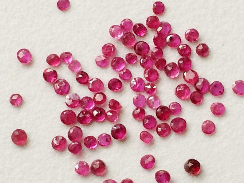 May include: A collection of small, faceted, round gemstones in varying shades of red and pink. The gemstones are scattered across a white surface, with light reflecting off their cut surfaces. These appear to be loose rubies.