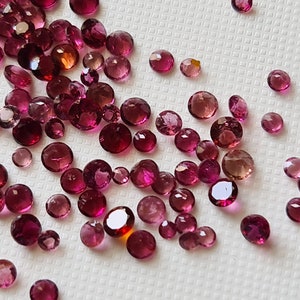 Op de afbeelding: Een verzameling ronde edelstenen in roze en rood, sommige met een lichte oranje tint, verspreid over een wit oppervlak.