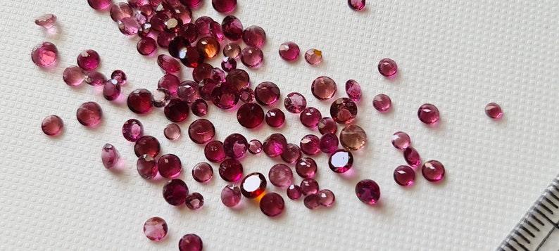 Op de afbeelding: Een verzameling ronde edelstenen in roze en rood, sommige met een lichte oranje tint, verspreid over een wit oppervlak.