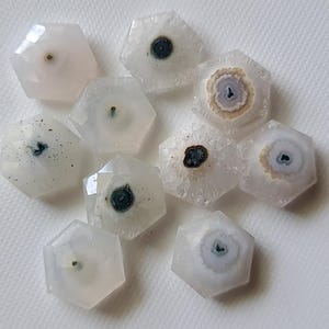Puede incluir: Diez cabujones de ágata hexagonales blancos con varios patrones de inclusiones negras, grises y marrones. Los cabujones están pulidos y tienen un acabado liso y brillante.