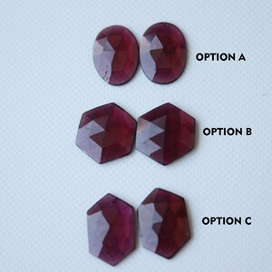 May include: Three sets of gemstone cabochons in different shapes. Option A is two oval cabochons, Option B is two hexagonal cabochons, and Option C is two oval cabochons. All cabochons are a deep reddish-purple color.