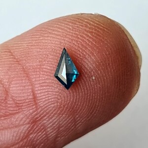 Op de afbeelding: Een kleine, vlieger-vormige, teal-kleurige edelsteen met een gefacetteerd oppervlak. De edelsteen wordt tussen twee vingers gehouden, wat de grootte en kleur laat zien. De slijping van de edelsteen creëert een spel van licht en schaduw.