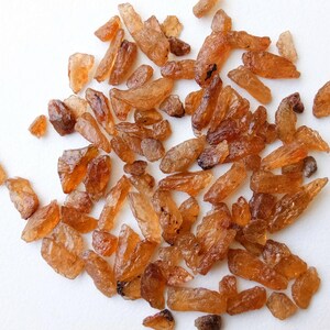 May include: A collection of small, brown, rough, and irregularly shaped gemstones. The gemstones are scattered on a white surface.