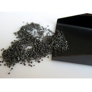 May include: A small black scoop with a pile of black, rough, granular material spilling out of it onto a white surface.