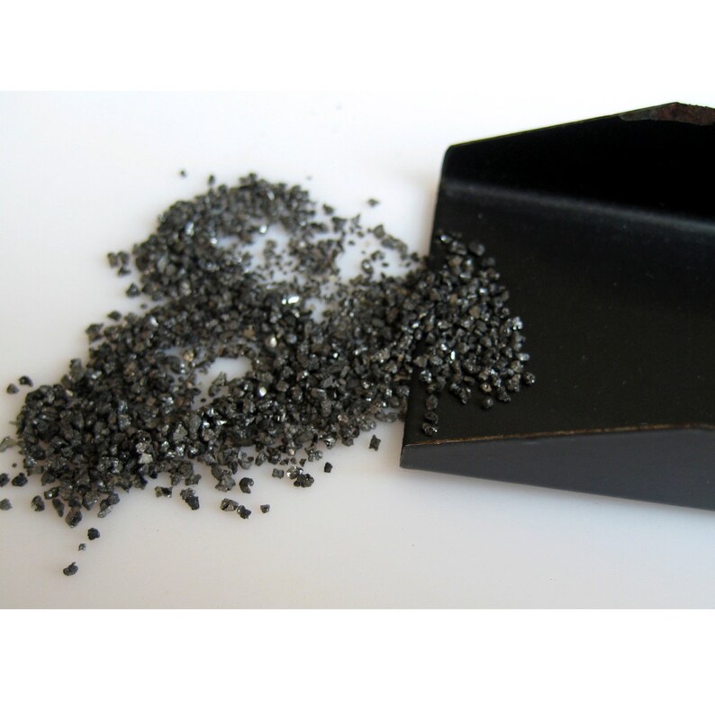 May include: A small black scoop with a pile of black, rough, granular material spilling out of it onto a white surface.