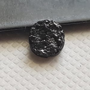 Könnte beinhalten: Eine schwarze, runde, strukturierte Münze mit einem erhabenen Design.