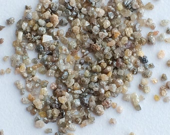 May include: A close-up view of a pile of small, rough, grey, brown, and clear diamonds. The diamonds are scattered on a white surface.
