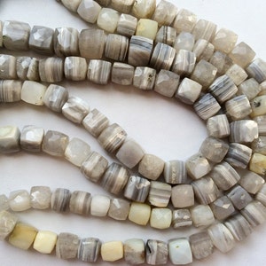 May include: A strand of gray and white agate beads with a faceted, square shape. The beads are strung on a cord and are ready to be used for jewelry making.