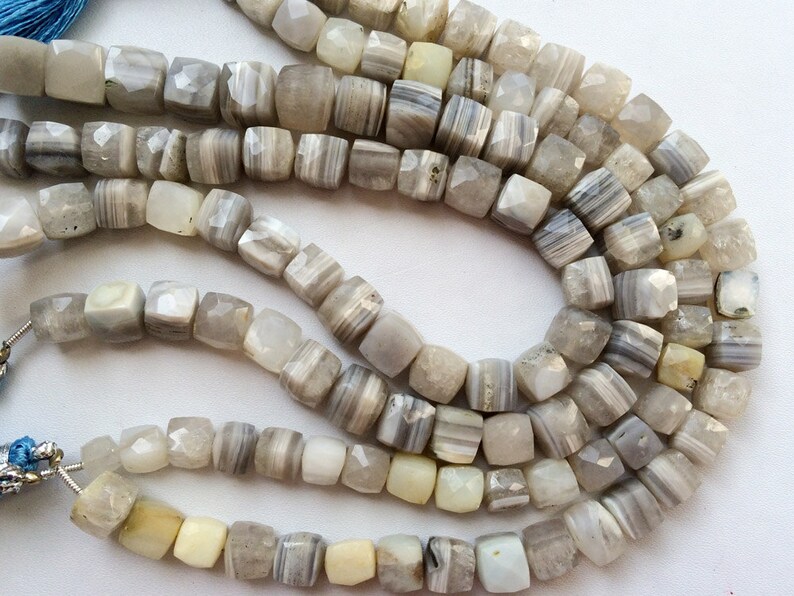May include: A strand of gray and white agate beads with a faceted, square shape. The beads are strung on a cord and are ready to be used for jewelry making.