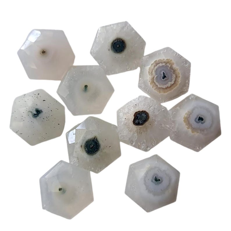 May include: Ten white agate gemstone cabochons with a variety of patterns in shades of gray and brown. The cabochons are cut into a hexagonal shape with a flat back and a domed top.