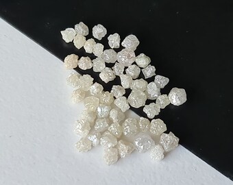 May include: A collection of small, white, rough diamonds. The diamonds are various shapes and sizes, and they are scattered on a white surface.