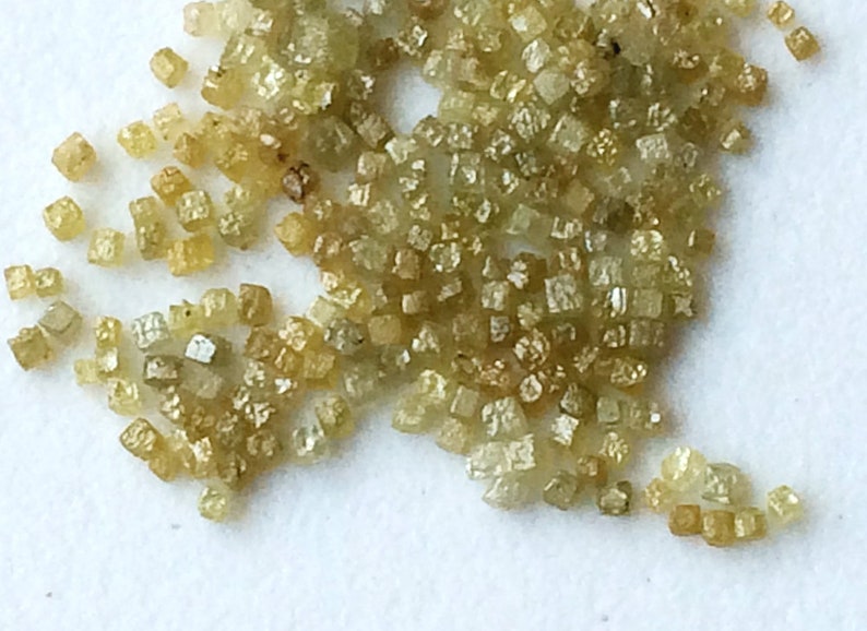 May include: A close-up of a pile of small, light green, faceted diamonds. The diamonds are scattered on a white surface.