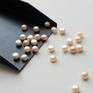 May include: A scoop of round, off-white pearls with a slight pink hue. The pearls are scattered on a white surface and spilling out of a black scoop.