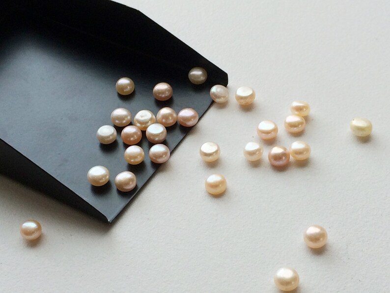 May include: A scoop of round, off-white pearls with a slight pink hue. The pearls are scattered on a white surface and spilling out of a black scoop.