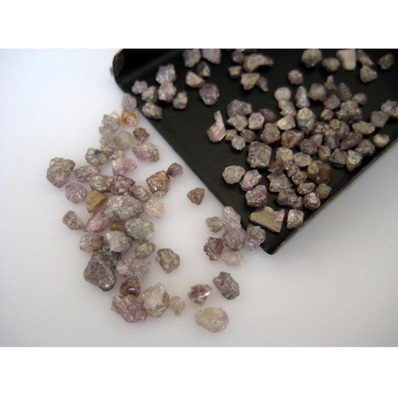 May include: A close-up of a pile of small, rough, pink and gray diamonds. The diamonds are scattered on a white surface and in a black tray.