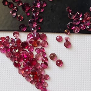 Op de afbeelding: Een verzameling roze en rode edelstenen, sommige met een lichte paarse tint, zijn verspreid over een wit oppervlak. De edelstenen zijn rond en hebben een gladde, gepolijste afwerking.