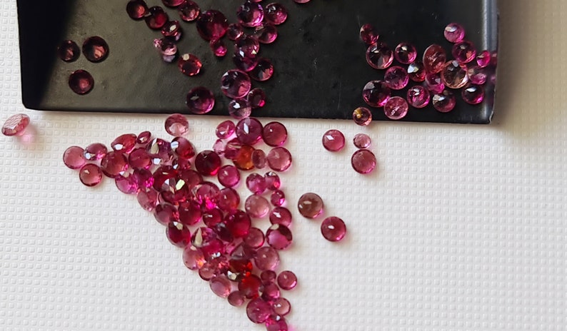 Op de afbeelding: Een verzameling roze en rode edelstenen, sommige met een lichte paarse tint, zijn verspreid over een wit oppervlak. De edelstenen zijn rond en hebben een gladde, gepolijste afwerking.