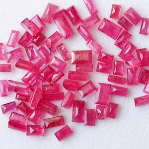 Puede incluir: Una colección de piedras preciosas rectangulares de color rosa, probablemente rubíes, esparcidas sobre una superficie blanca. Las piedras preciosas son de varios tamaños y formas, algunas tienen una forma rectangular más alargada.