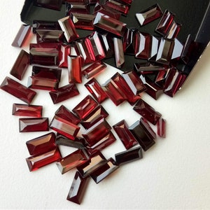 May include: A collection of red, rectangular-shaped gemstones, scattered on a white surface. The gemstones are faceted and have a glossy finish.