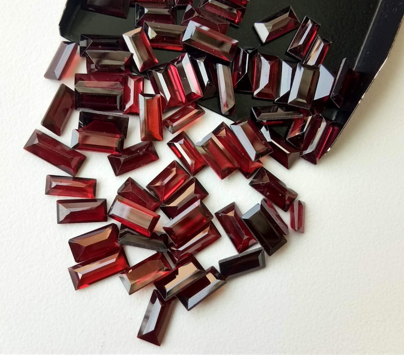 May include: A collection of red, rectangular-shaped gemstones, scattered on a white surface. The gemstones are faceted and have a glossy finish.
