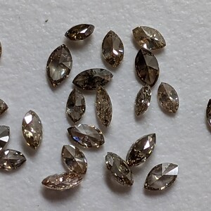 Puede incluir: Una colecci&oacute;n de 20 diamantes marrones en forma de marquesa, esparcidos sobre una superficie blanca. Los diamantes est&aacute;n facetados y tienen un ligero brillo.