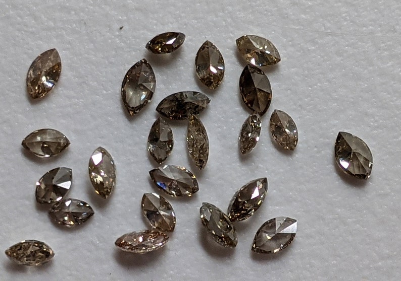 Puede incluir: Una colecci&oacute;n de 20 diamantes marrones en forma de marquesa, esparcidos sobre una superficie blanca. Los diamantes est&aacute;n facetados y tienen un ligero brillo.