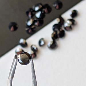 May include: A single black gemstone held in tweezers. The gemstone is round and faceted. There are other black gemstones scattered on a white surface.