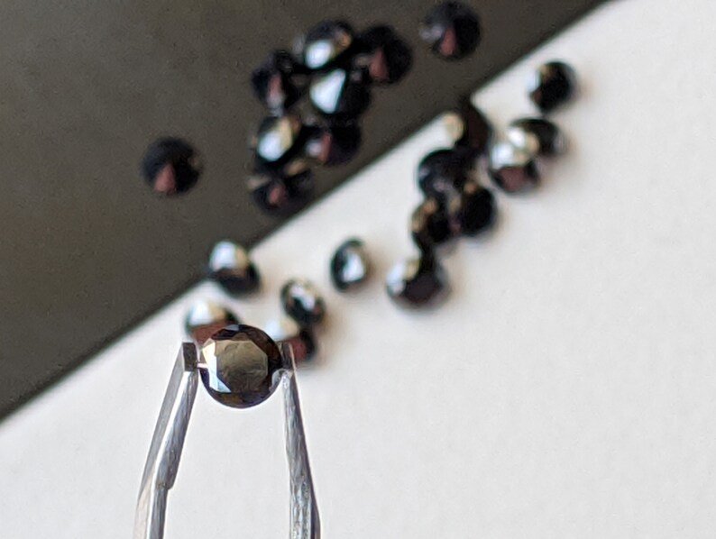 May include: A single black gemstone held in tweezers. The gemstone is round and faceted. There are other black gemstones scattered on a white surface.