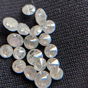 May include: A close-up of a pile of small, round, clear, faceted diamonds. The diamonds are scattered on a dark, textured surface.
