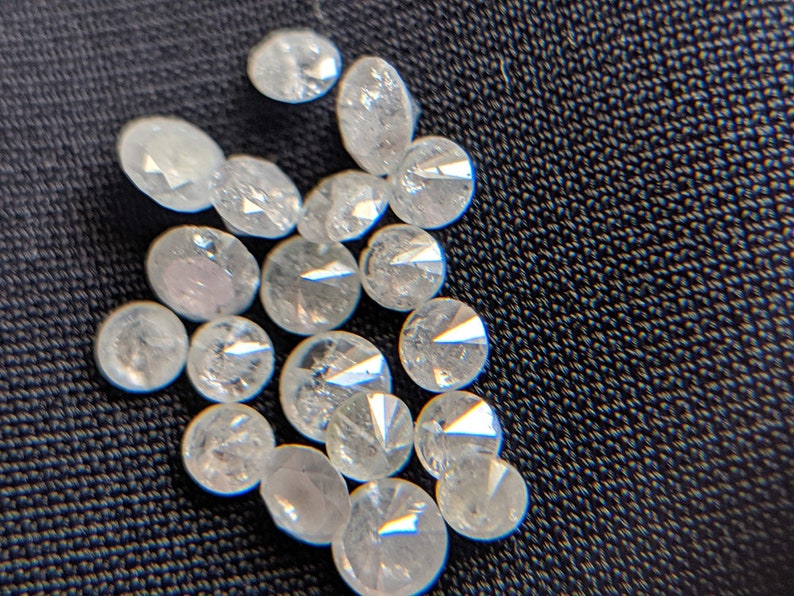 May include: A close-up of a pile of small, round, clear, faceted diamonds. The diamonds are scattered on a dark, textured surface.