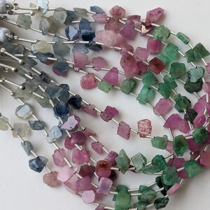 Può includere: Un filo di pietre preziose grezze e sfaccettate in tonalità di blu, rosa e verde. Le pietre sono infilate su un filo d'argento e hanno un aspetto naturale e grezzo.