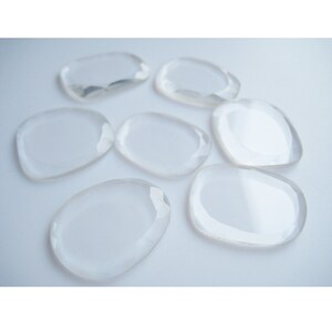 May include: Eight clear, smooth, and polished crystal stones in various shapes and sizes. The stones are arranged on a white surface.