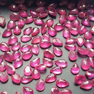 May include: A collection of pear-shaped pink gemstones, likely rubies, displayed on a black surface. The gemstones are faceted and have a high polish.