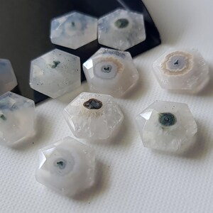May include: Eight white, hexagonal, agate cabochons with green and brown inclusions. The cabochons are polished and have a smooth, glossy finish.