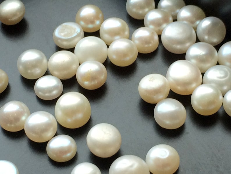 May include: A collection of white round pearls of various sizes, scattered on a dark surface.