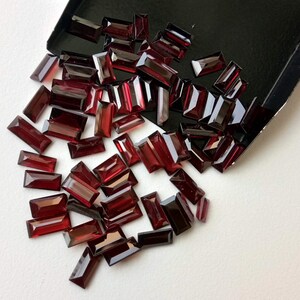 May include: A large pile of dark red rectangular gemstones, scattered on a white surface. The gemstones are all the same size and shape, and they have a glossy finish.