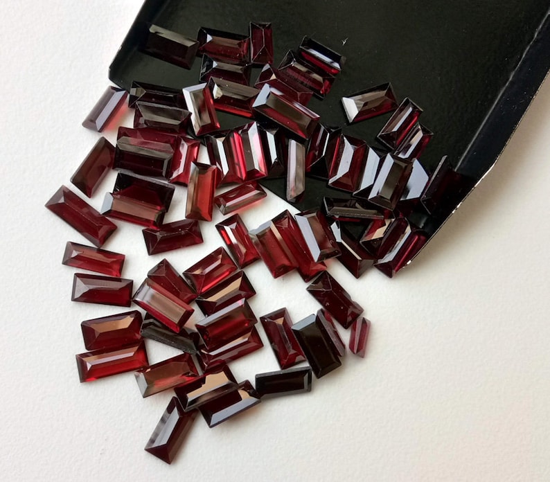 May include: A large pile of dark red rectangular gemstones, scattered on a white surface. The gemstones are all the same size and shape, and they have a glossy finish.