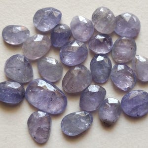 May include: A collection of loose, faceted tanzanite gemstones in varying shades of light purple and blue. Each stone features a polished, irregular oval shape with multiple flat facets that reflect light across their translucent surfaces.
