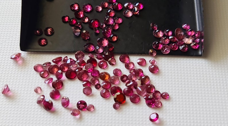 Op de afbeelding: Een verzameling losse roze edelstenen, sommige in een zwarte schaal en andere verspreid over een wit oppervlak.